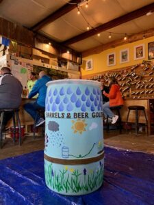 a blue rain barrel is seen, it has been painted with raindrops, a sun, clouds, and flowers and says "Barrels & Beer Golden..."